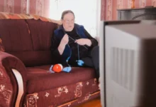 stará žena s překvapeným výrazem z gauče sleduje televizi zatímco plete