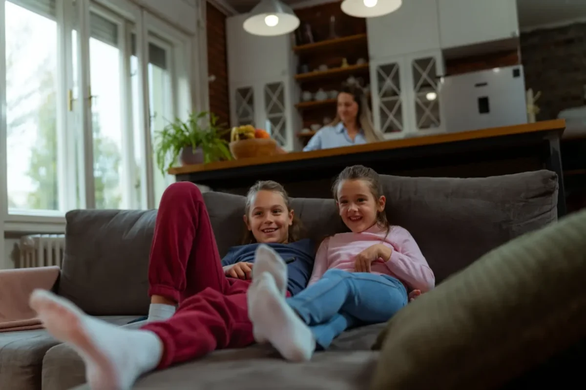 dvě malé sestry spokojeně sledují televizi z gauče, zatímco se jejich matka usmívá v pozadí