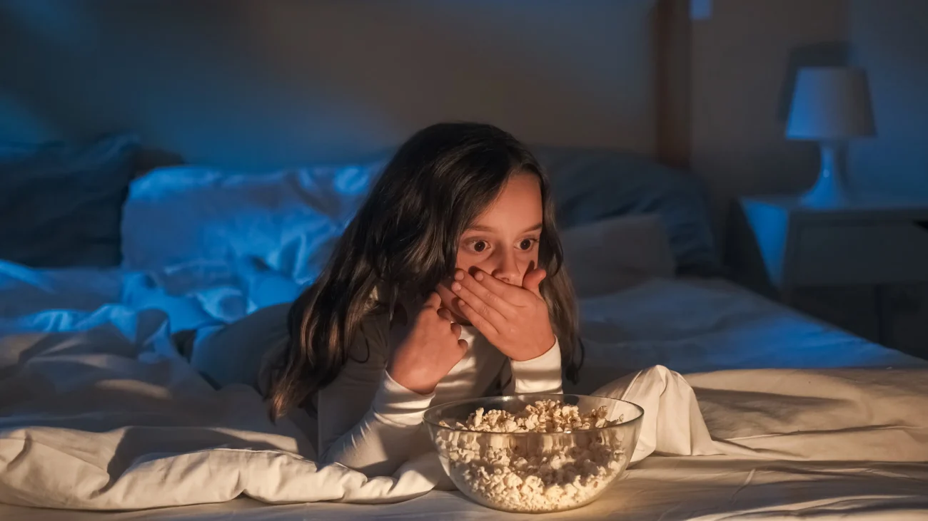 shutterstock_2533061253-2 | TVGURU.cz Holčička vyděšená leží s popcornem na posteli.