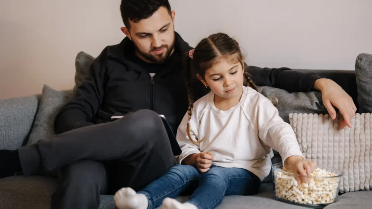 Muž s dcerou sedí na gauči a nabírají si popcorn.