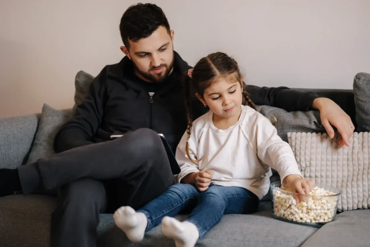Muž s dcerou sedí na gauči a nabírají si popcorn.