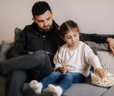 Muž s dcerou sedí na gauči a nabírají si popcorn.