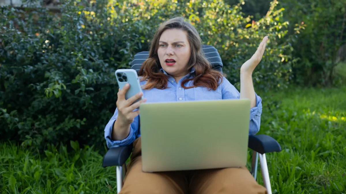 Žena sedí venku a nevěřícně kouká do telefonu.