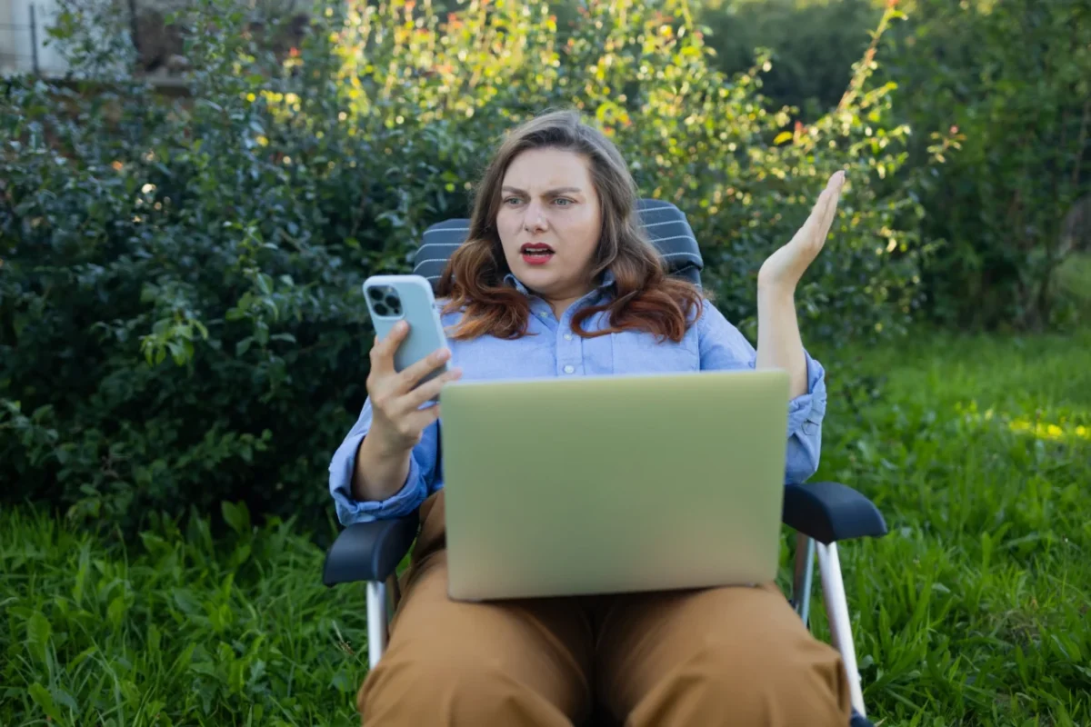 Žena sedí venku a nevěřícně kouká do telefonu.