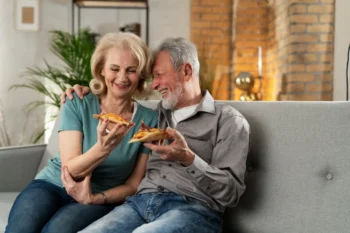 Starší pár sedí na gauči a dává si pizzu.