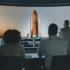 Vývojáři u počítačů a s plátnem s raketou.