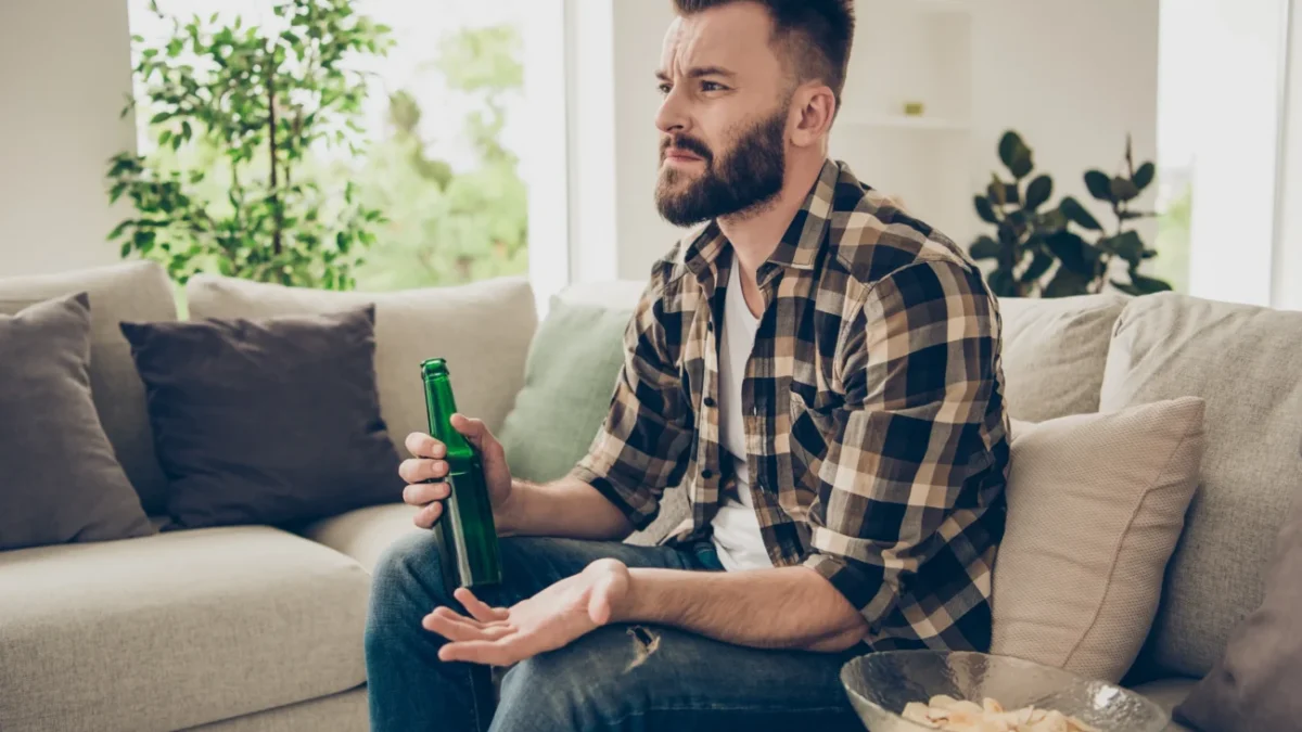 Muž v ruce drží pivo a nechápavě kouká na televizi.