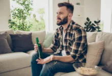 Muž v ruce drží pivo a nechápavě kouká na televizi.