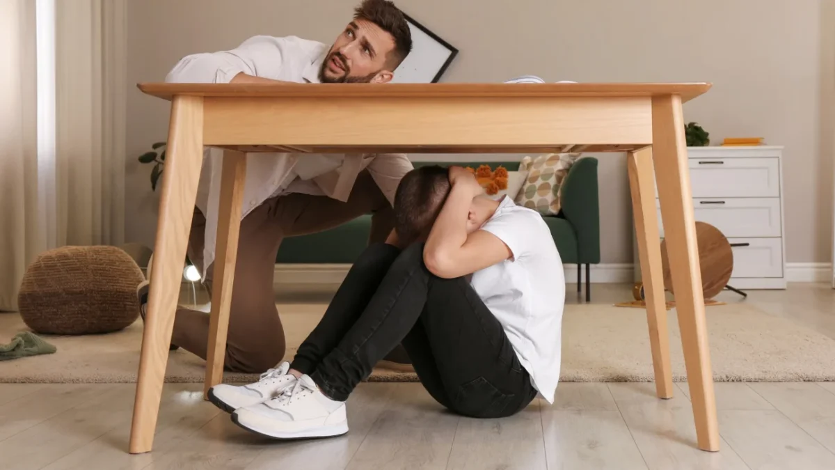 Chlapeček se schovává pod stolem a muž se ho snaží vylákat ven.