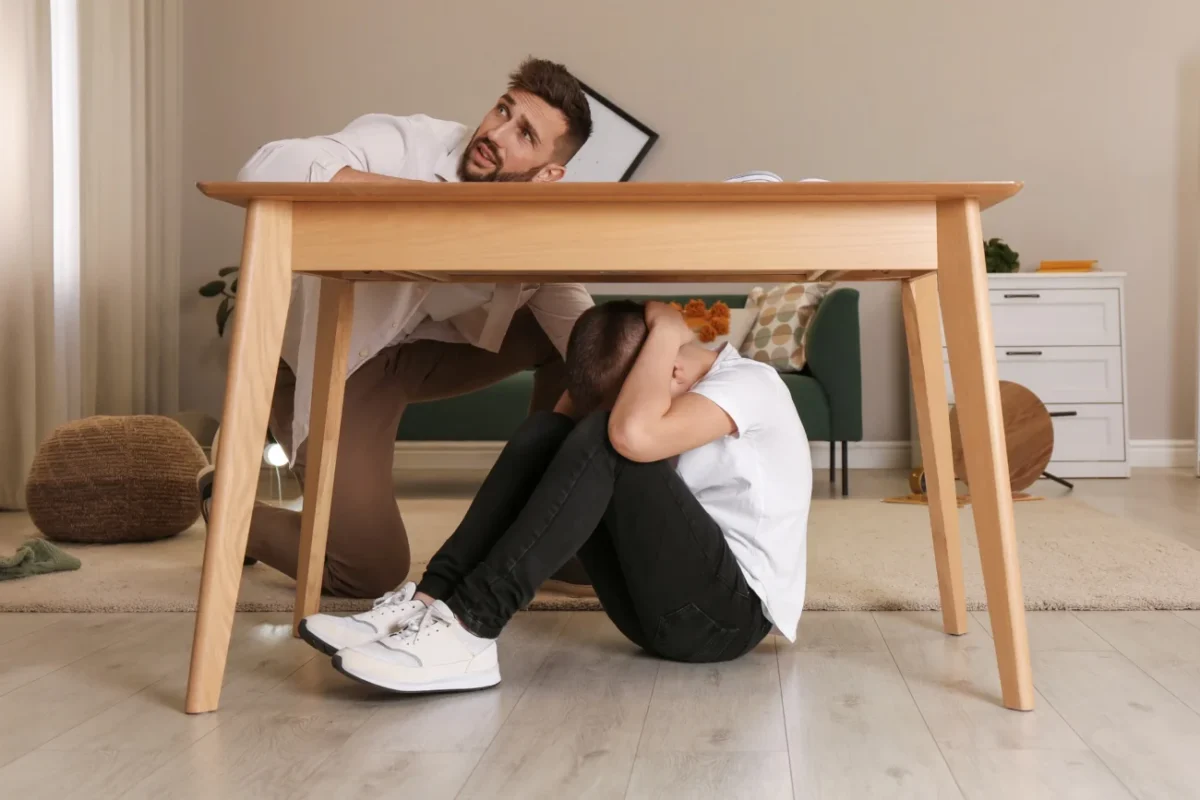 Chlapeček se schovává pod stolem a muž se ho snaží vylákat ven.