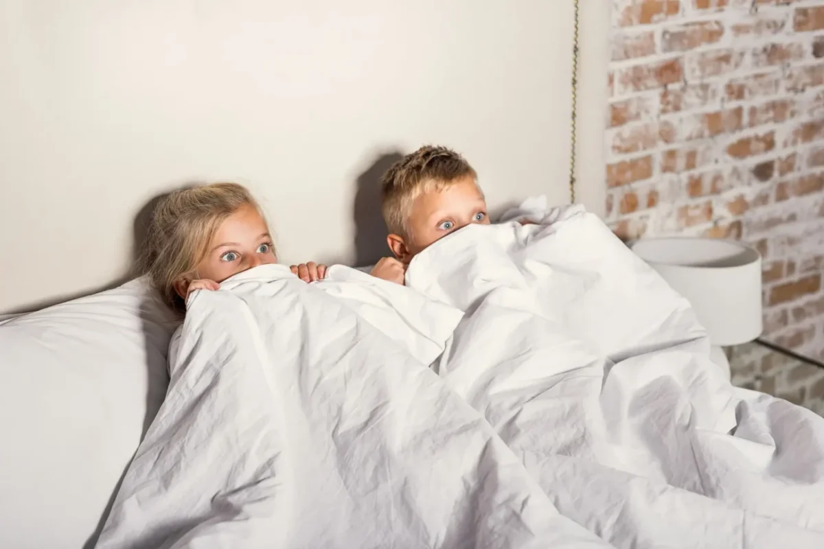 dvě děti pod peřinou vyděšeně sledují televizi z postele