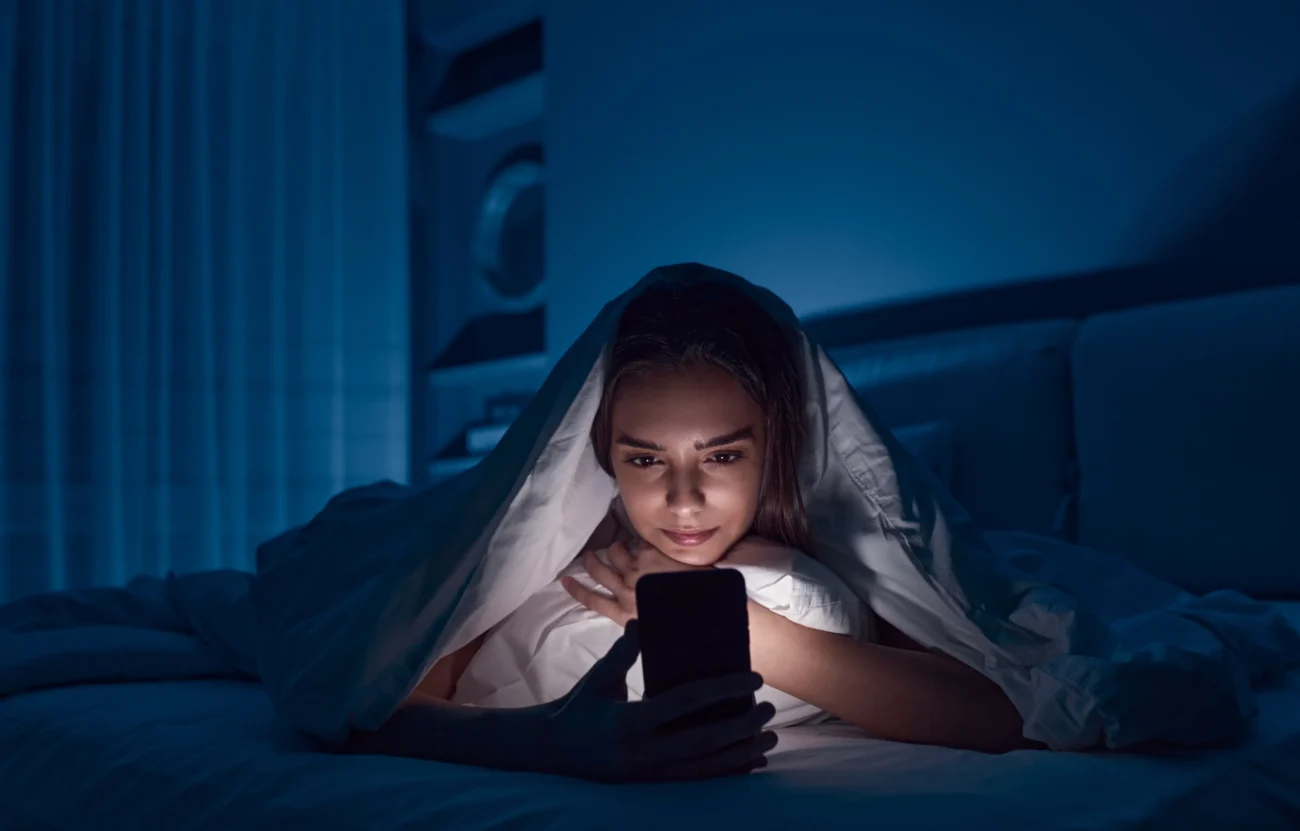shutterstock_2109469025-2 | TVGURU.cz Žena leží v posteli pod peřinou a je na telefonu.