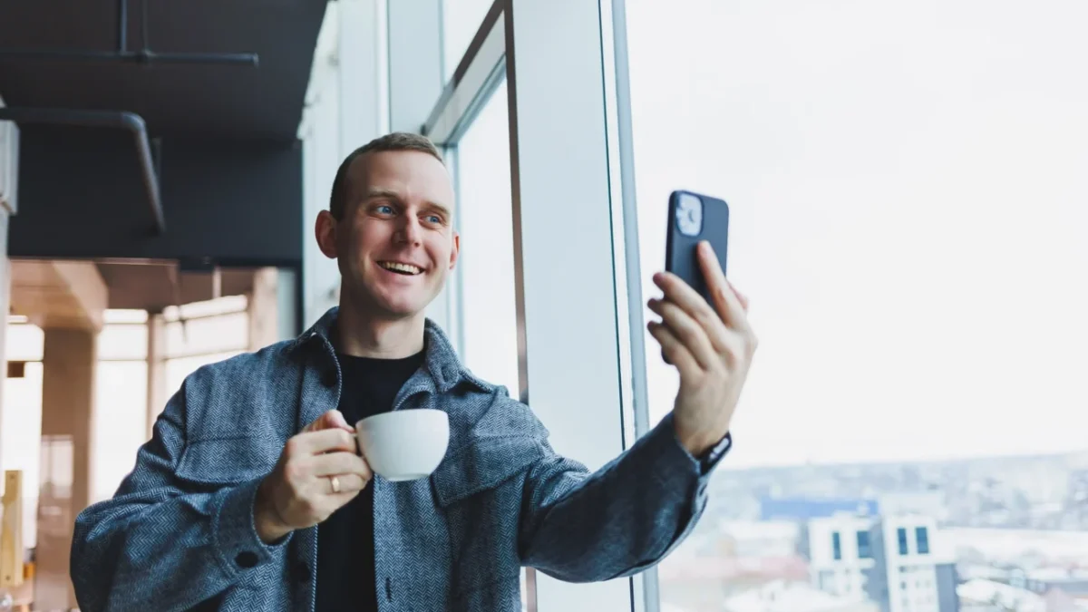 Muž se usmívá, drží v ruce kávu a kouká se do telefonu.