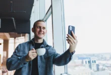 Muž se usmívá, drží v ruce kávu a kouká se do telefonu.