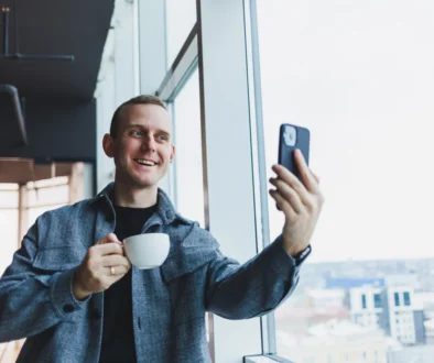 Muž se usmívá, drží v ruce kávu a kouká se do telefonu.