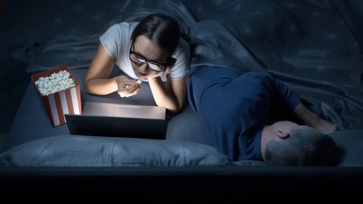 Žena v noci kouká v posteli na film a jí popcorn.