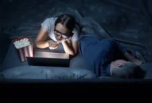 Žena v noci kouká v posteli na film a jí popcorn.