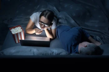 Žena v noci kouká v posteli na film a jí popcorn.