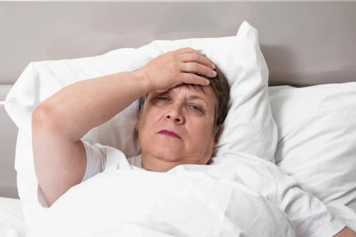 Babička leží v posteli a chytá se za čelo.