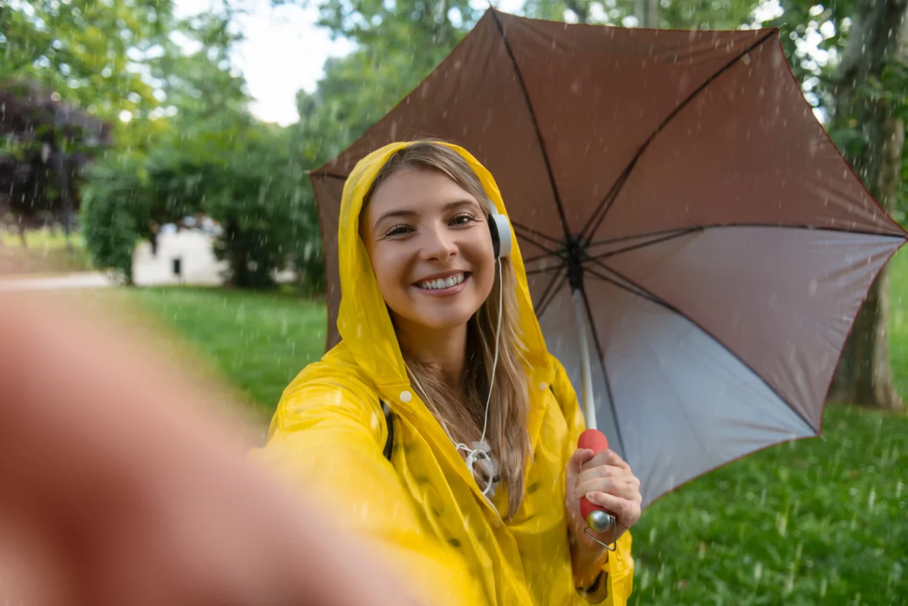 shutterstock_2434553319-2 | TVGURU.cz Žena oblečená v pláštěnce si pod deštníkem fotí selfie.