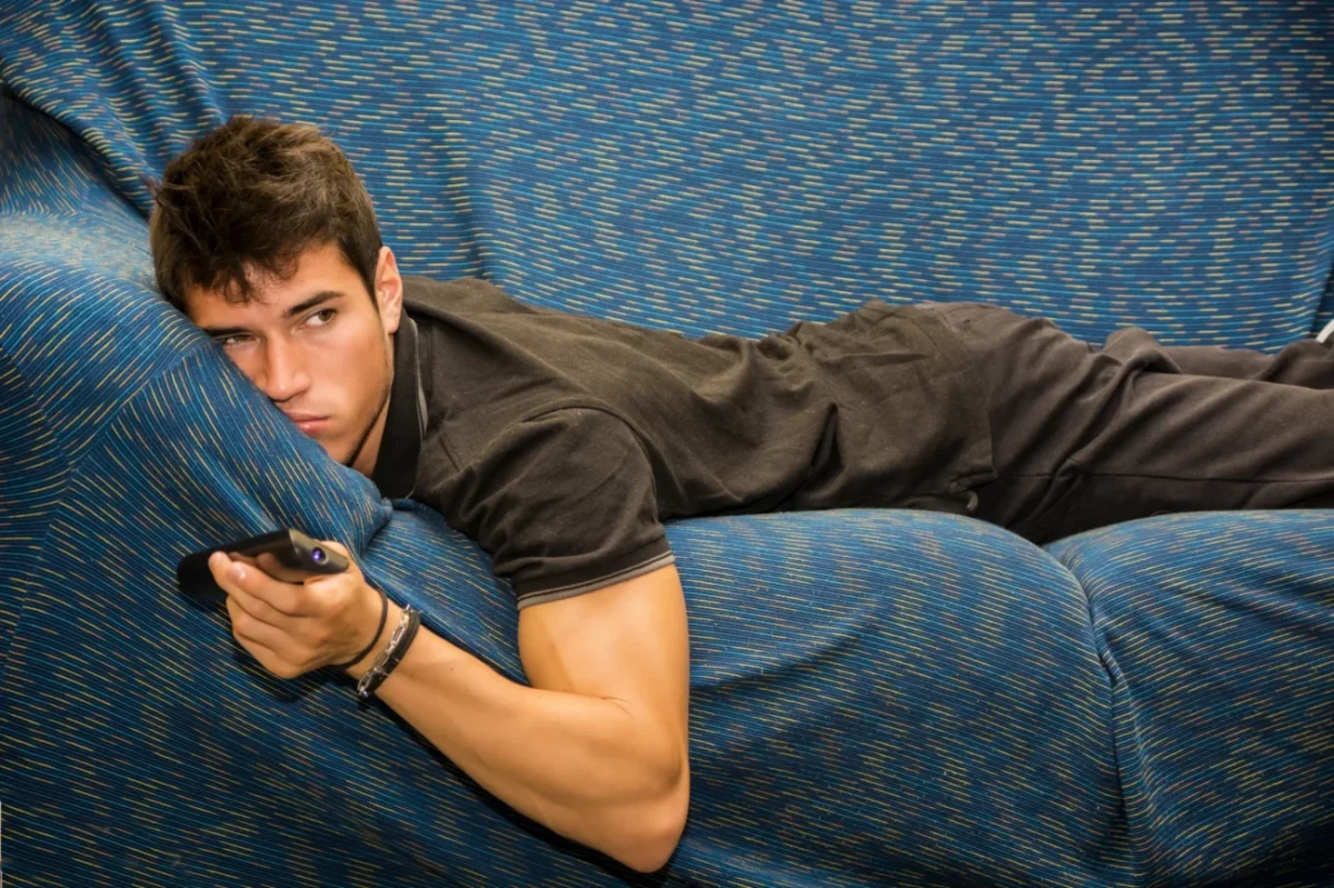 Muž leží otrávený na pohovce a má v ruce ovladač od TV.