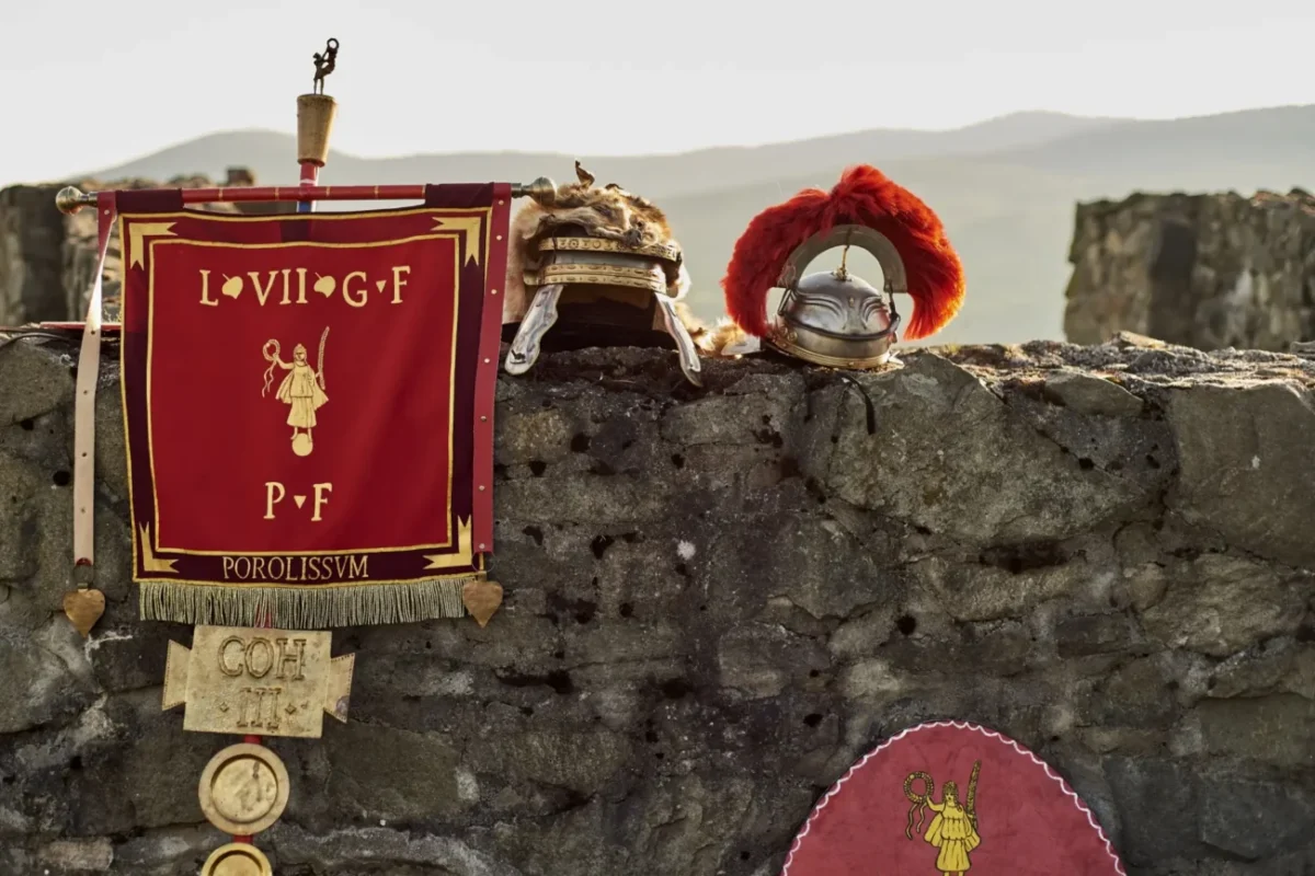 vlajka římské legie opřená o kamennou stěnu, na které je římská přilbice