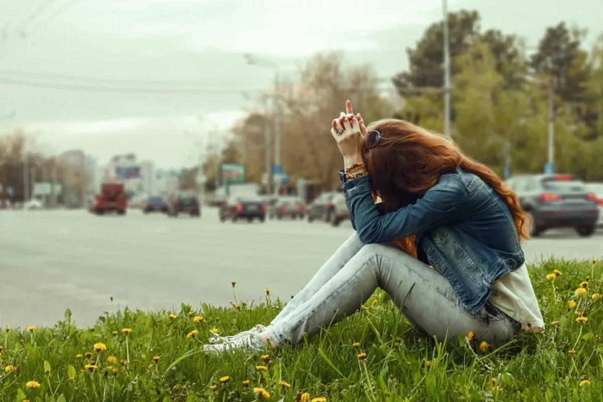 Žena sedí venku a hlavu má složenou mezi nohama.