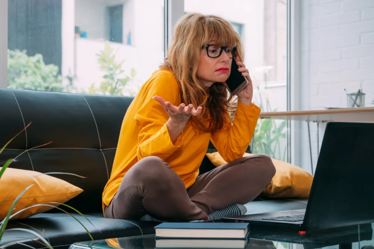 Žena sedí na gauči, telefonuje a tváří se šokovaně.