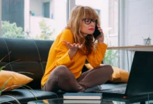 Žena sedí na gauči, telefonuje a tváří se šokovaně.