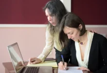 Jedna žena něco vyplňuje na papíře a druhá na něco kouká v notebooku.
