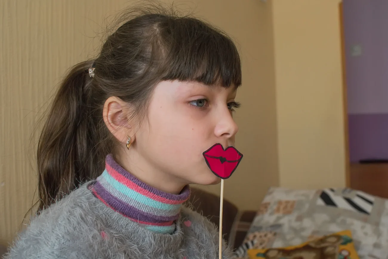 Holčička má před obličejem na špejli papírek ve tvaru pusy.