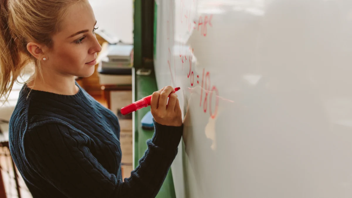 Mladá blond žena s culíkem - studentka. Píše markerem na bílou tabuli. Z profilu. Matematický příklad.