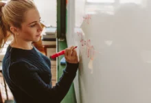 Mladá blond žena s culíkem - studentka. Píše markerem na bílou tabuli. Z profilu. Matematický příklad.