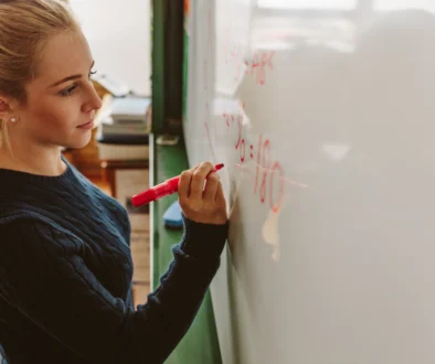 Mladá blond žena s culíkem - studentka. Píše markerem na bílou tabuli. Z profilu. Matematický příklad.