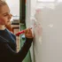 Mladá blond žena s culíkem - studentka. Píše markerem na bílou tabuli. Z profilu. Matematický příklad.