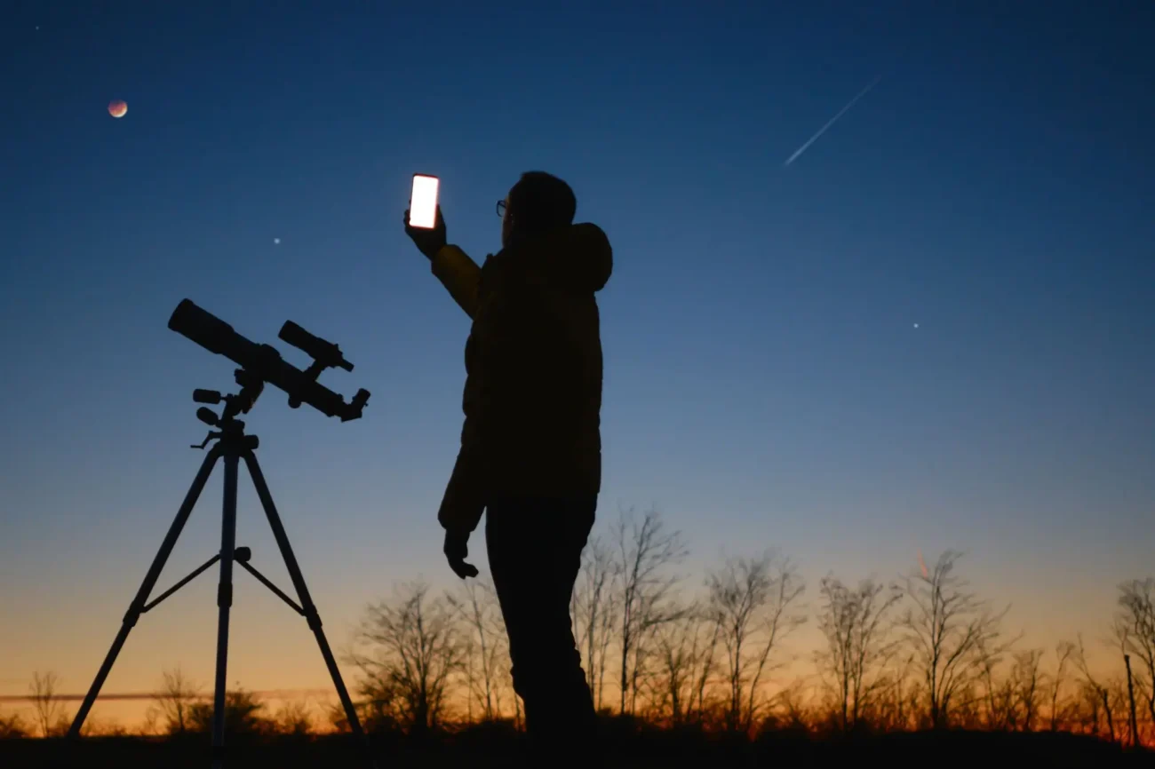 Muž který se dívá na zatmění měsíce, má před sebou teleskop a fotí si to mobilem