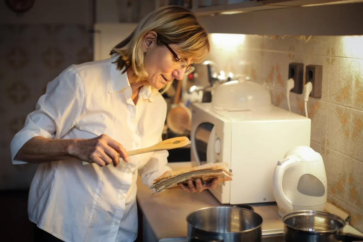 Žena středního věku večer v kuchyni drží v rukou vařečku starý sešit s recepty