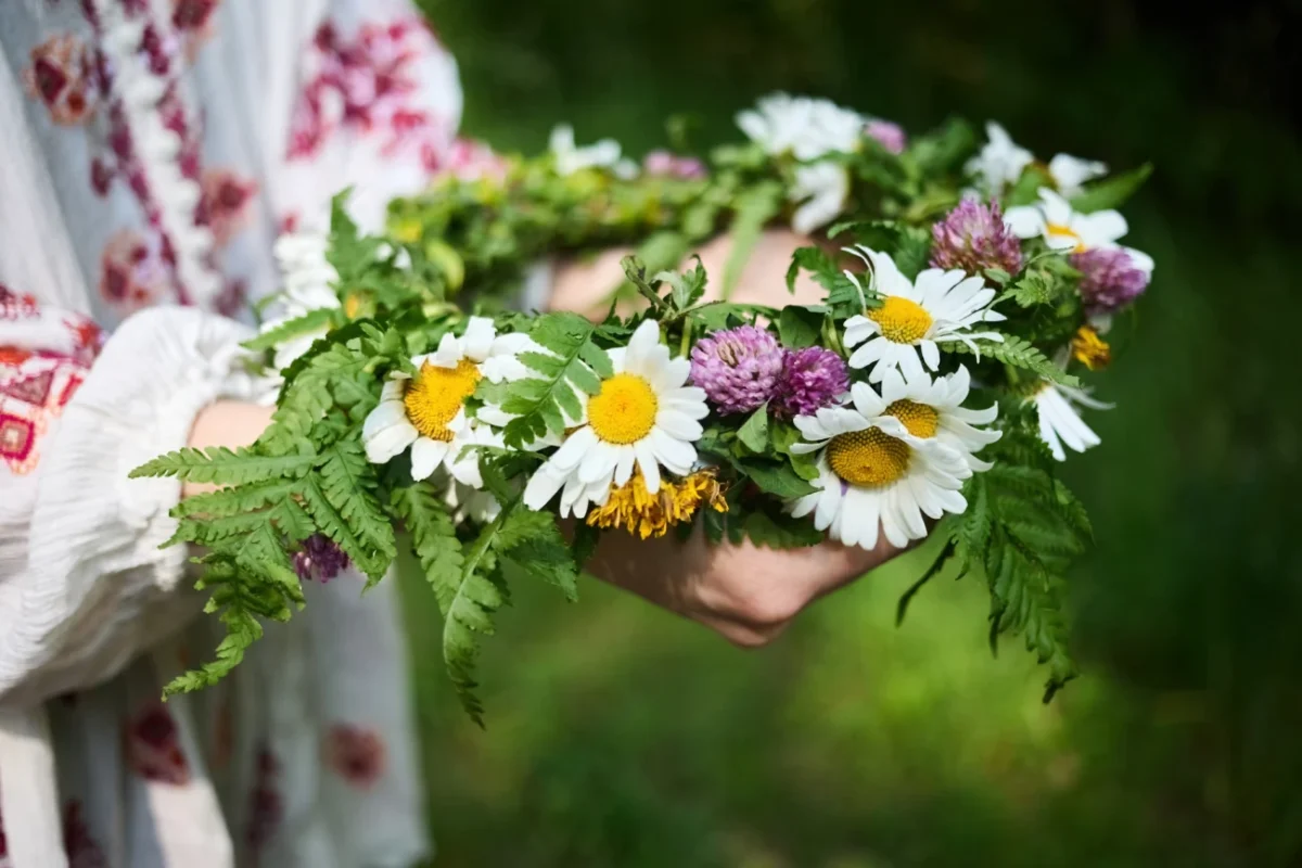 detail věnce z lučního kvítí v rukou oděných do kroje (vyšívané bílé rukávce). Zelená louka/tráva na pozadí