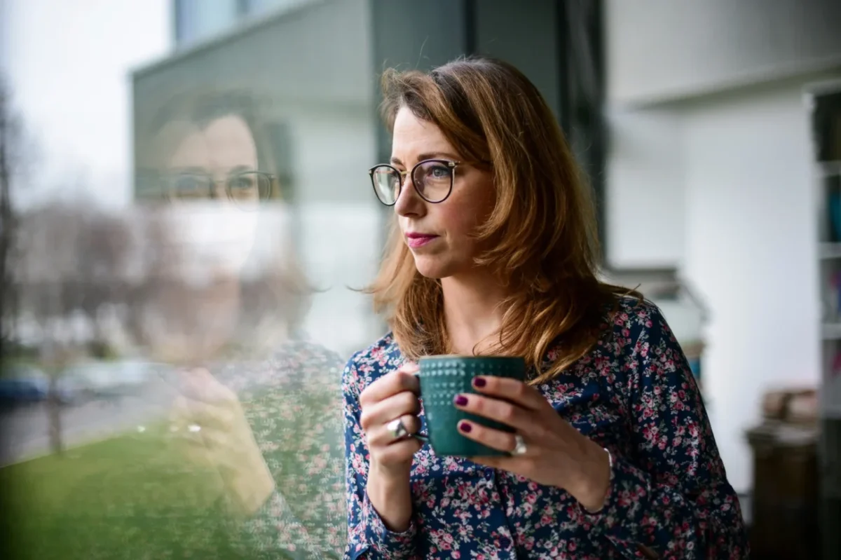 Žena dívající se z okna s hrnkem čaje.