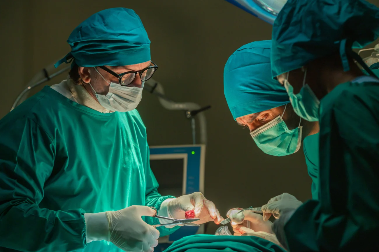 shutterstock_2275814879 | TVGURU.cz Detail - trojice chirurgů soustředěně operuje (zelené oblečení, modré pokrývky hlavy)