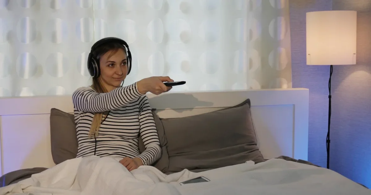 Žena leží v posteli, má sluchátka na uších a míří ovladačem.