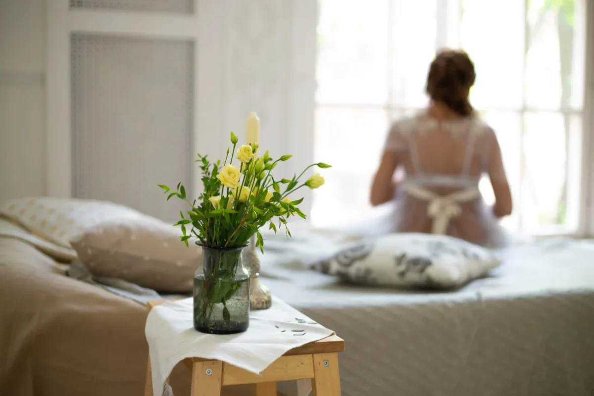 Žena vstává z postele a před ní na stolečku stojí kytice.