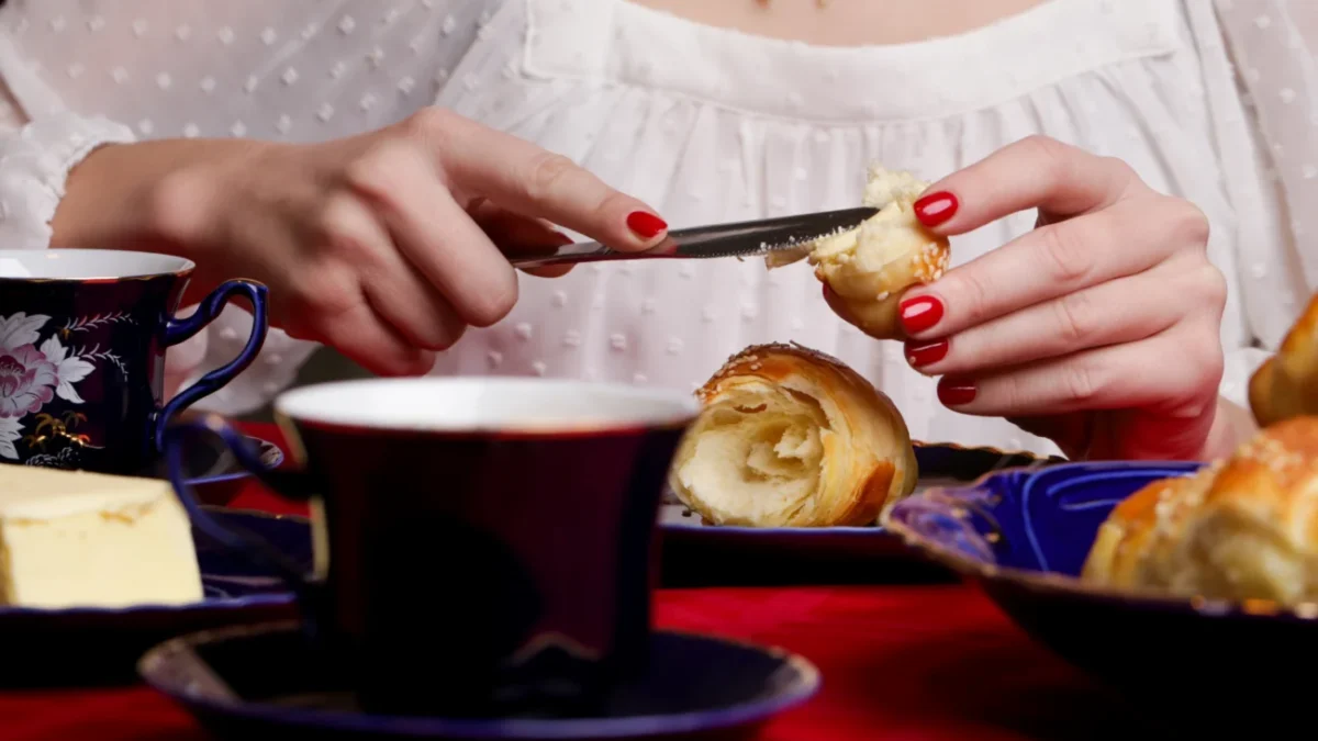 žena v bílé krajové halence si maže máslem croissant, zřejmě snídá. hlava ženy je mimo záběr - detail na její snídani = stůl s jídlem