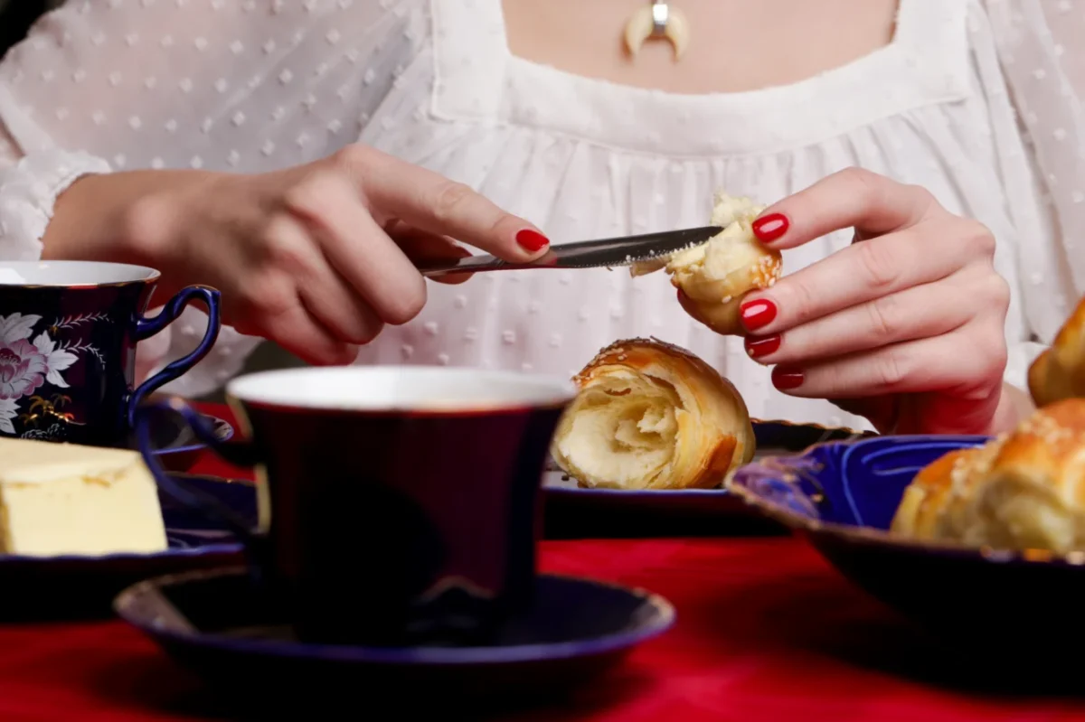 žena v bílé krajové halence si maže máslem croissant, zřejmě snídá. hlava ženy je mimo záběr - detail na její snídani = stůl s jídlem