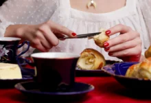žena v bílé krajové halence si maže máslem croissant, zřejmě snídá. hlava ženy je mimo záběr - detail na její snídani = stůl s jídlem