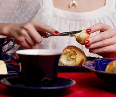 žena v bílé krajové halence si maže máslem croissant, zřejmě snídá. hlava ženy je mimo záběr - detail na její snídani = stůl s jídlem