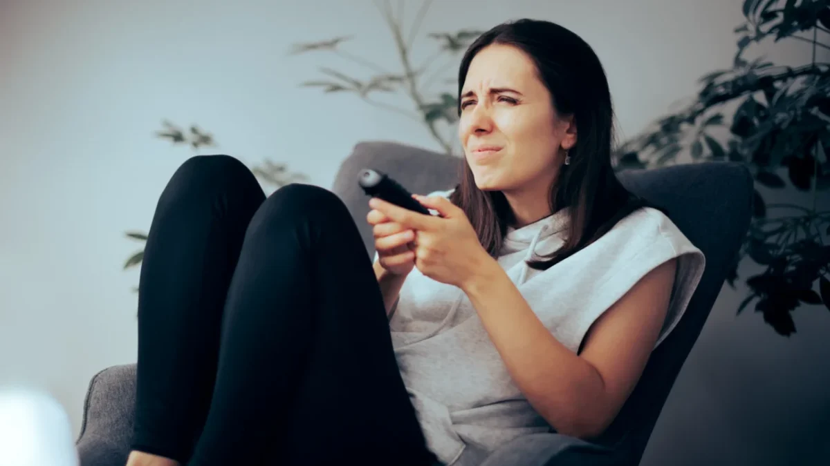 Žena sedí na křesle a nechápavě se dívá na televizi.