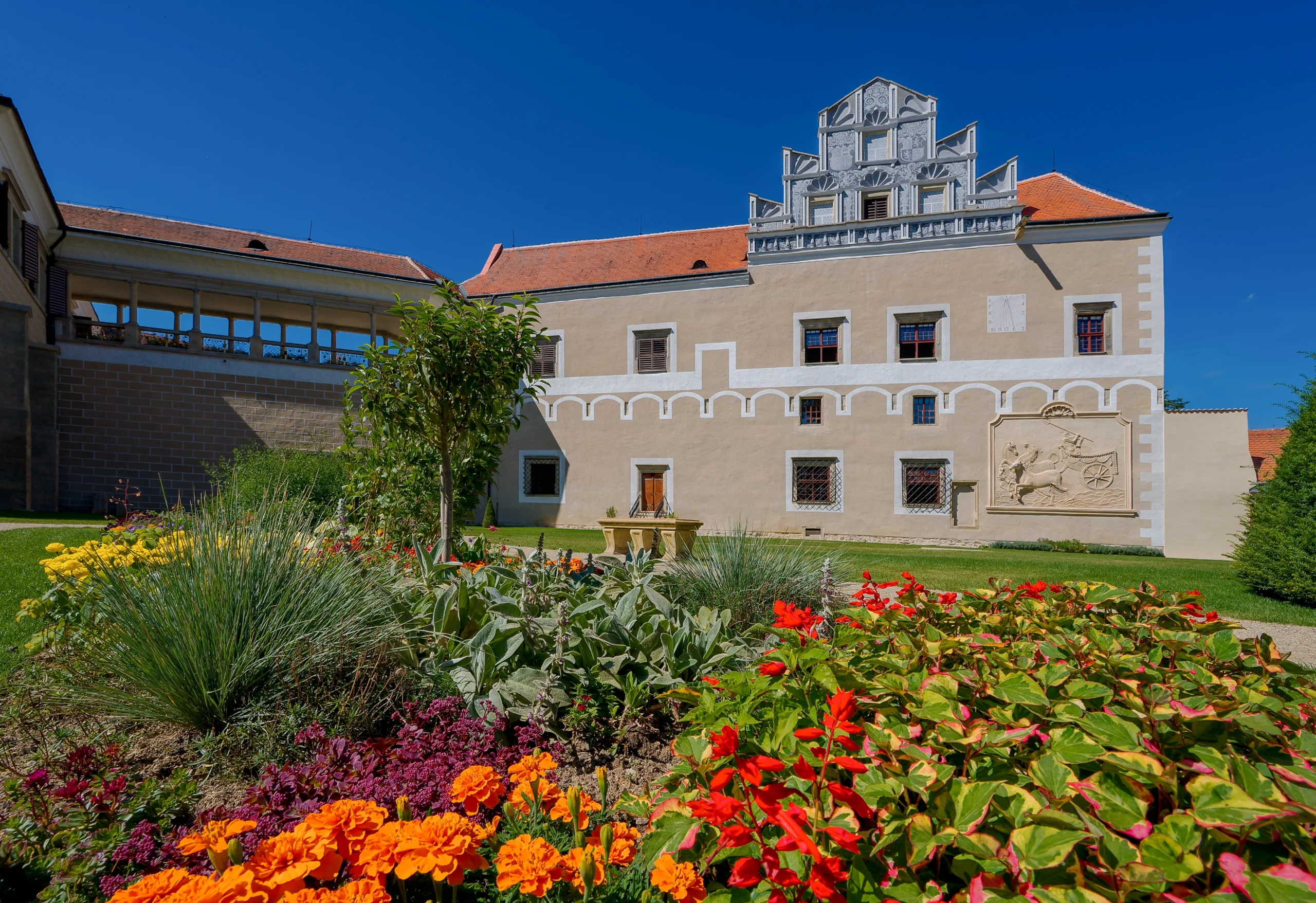 shutterstock_2506324241 | TVGURU.cz Zámecká zahrada - zámek Telč v pozadí, barevné květinové záhony v popředí. Krásný jasný slunný den, modrá obloha