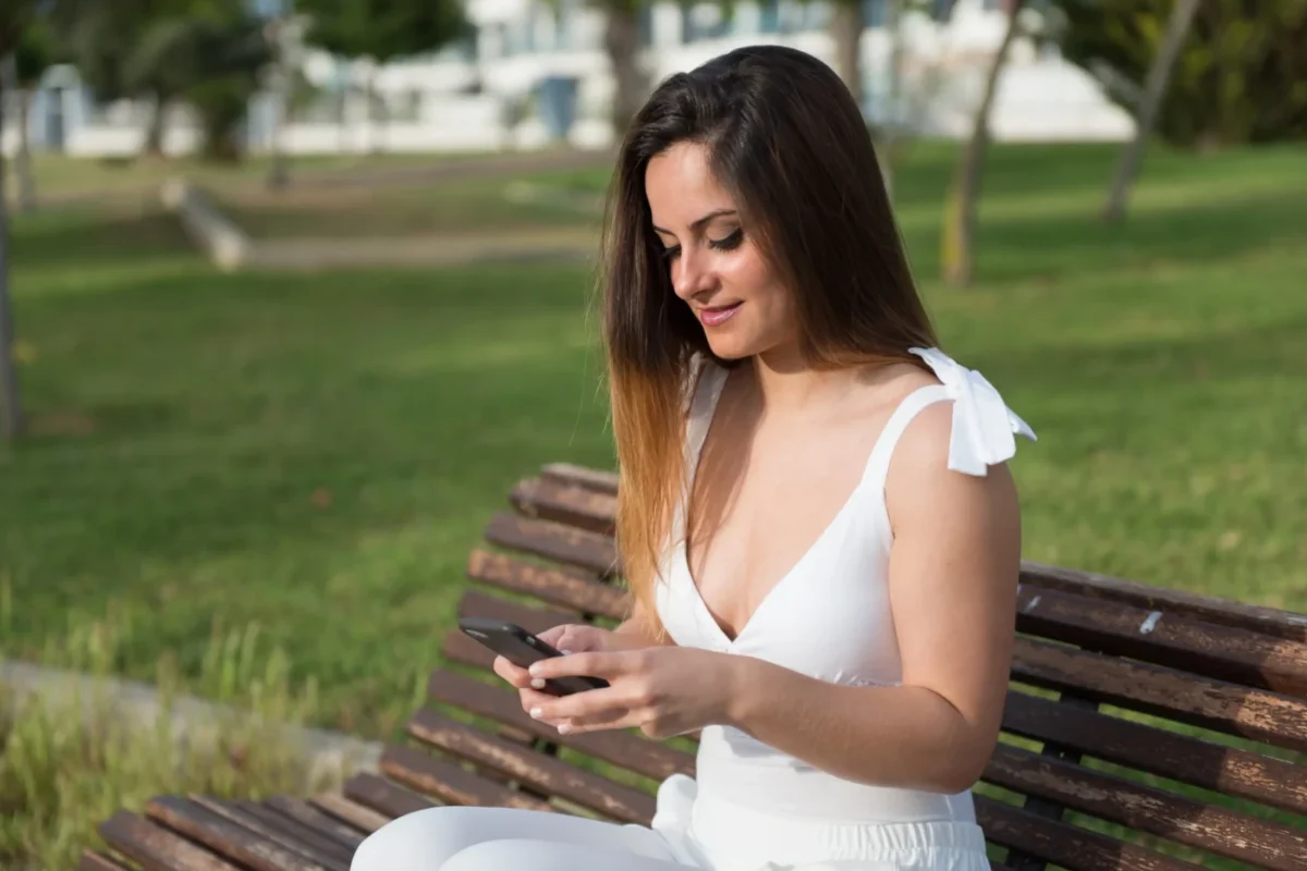 Žena sedí na lavičce a usmívá se do telefonu.