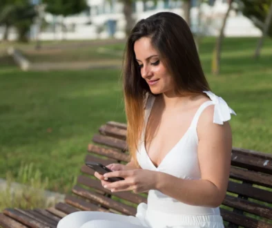 Žena sedí na lavičce a usmívá se do telefonu.